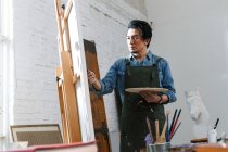 Концентрированный молодой азиатский художник в фартуке держит палитру и живопись картины в студии — стоковое фото