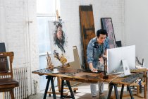 Joven artista masculino sosteniendo la paleta y utilizando la computadora de escritorio en el estudio - foto de stock