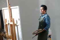 Concentrated asian artist holding palette and painting picture in studio — Stock Photo