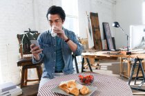 Молодой азиат пьет молоко и пользуется смартфоном во время завтрака в арт-студии — стоковое фото