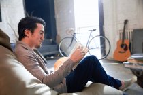 Vista lateral de sorrir jovem asiático homem sentado no feijão saco cadeira e leitura livro em casa — Fotografia de Stock