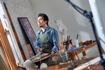 Low angle view of thoughtful male painter holding palette and looking at easel in studio — Stock Photo