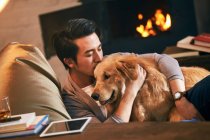 Joven guapo sentado en la silla de la bolsa de frijol y abrazando a su perro en casa - foto de stock