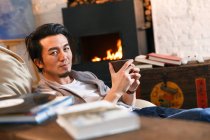 Guapo asiático hombre celebración taza con Caliente bebida y mirando cámara en casa - foto de stock