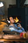 Pensive asian man in headphones holding cup with hot beverage and looking away at home — Stock Photo