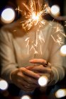 Tiro cortado de pessoa segurando queimando sparkler no fundo festivo borrado — Fotografia de Stock