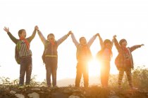 Счастливые сельские ученики держатся и поднимают руки, стоя на холме на рассвете — стоковое фото