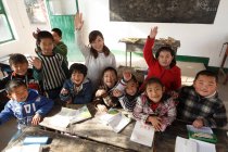 High angle view of rural female teacher and chinese pupils raising hands and smiling at camera in classroom — Stock Photo
