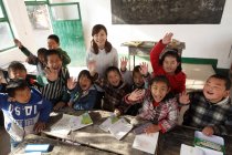 Высокий угол обзора сельских учительниц и китайских учеников, поднимающих руки и улыбающихся перед камерой в классе — стоковое фото