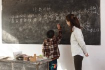 Young female chinese teacher looking at elementary school student writing on chalkboard — Stock Photo