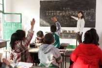 Rural female teacher pointing at chalkboard and chinese pupils raising hands in the classroom — Stock Photo
