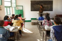Landlehrerin steht neben Tafel und chinesische Schüler sitzen an Schreibtischen im Klassenzimmer — Stockfoto