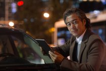 Зрілий азіатський чоловік стоїть біля машини і використовує цифровий планшет в нічному місті — стокове фото