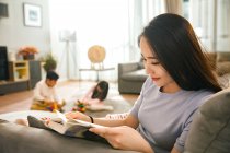 Smiling young woman reading book while children playing behind on carpet — Stock Photo