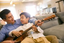 Feliz pai e filho chinês tocando guitarra acústica juntos em casa — Fotografia de Stock