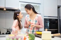Счастливые мама и дочь улыбаются друг другу, готовя вместе на кухне — стоковое фото
