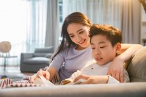 Smiling young mother with cute son sitting on couch and reading book together at home — Stock Photo