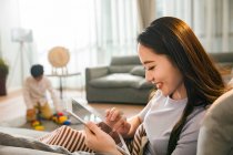 Side view of smiling woman using digital tablet while son playing with toys behind at home — Stock Photo