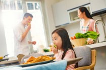 Adorable child eating apple while parents standing behind in kitchen — Stock Photo