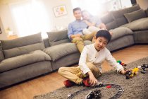 Happy parents sitting on couch and looking at smiling boy playing with toys on carpet — Stock Photo