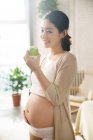 Smiling young pregnant woman holding green apple and standing at home — Stock Photo