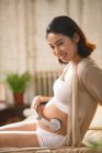 Side view of smiling young pregnant woman sitting on bed and holding headphones on belly — Stock Photo