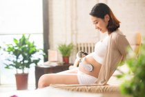 Vue latérale de la jeune femme enceinte souriante assise sur le lit et tenant des écouteurs sur le ventre — Photo de stock