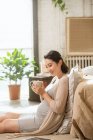 Seitenansicht einer lächelnden jungen Schwangeren, die zu Hause eine Tasse mit Heißgetränk hält — Stockfoto