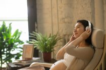 Vista lateral de mujer embarazada sonriente escuchando música en auriculares en casa - foto de stock