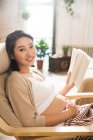 Feliz joven embarazada sosteniendo libro y sonriendo a la cámara en casa - foto de stock