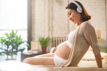 Вид сбоку улыбающейся беременной женщины, слушающей музыку в наушниках дома — стоковое фото