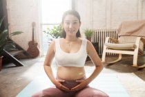 Счастливая молодая беременная женщина сидит на ковриках для йоги, трогает живот и улыбается в камеру дома — стоковое фото
