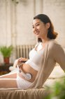 Усміхнена молода вагітна жінка сидить на ліжку і тримає навушники на животі — стокове фото