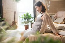 Side view of smiling young pregnant woman sitting on bed and touching belly — Stock Photo
