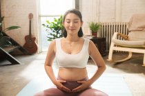 Улыбающаяся молодая беременная женщина сидит на ковриках для йоги и медитирует с закрытыми глазами дома — стоковое фото