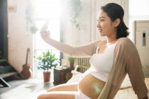 Vue latérale de la jeune femme enceinte souriante prenant selfie avec smartphone à la maison — Photo de stock