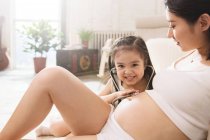 Adorable smiling child holding stethoscope and listening to belly of pregnant mother — Stock Photo