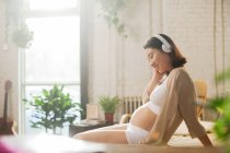 Вид сбоку улыбающейся молодой беременной женщины, слушающей музыку в наушниках дома — стоковое фото