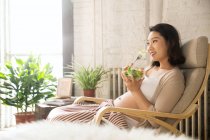 Side view of smiling young pregnant woman sitting in armchair and eating vegetable salad at home — Stock Photo