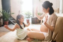 Вид сбоку на счастливого ребенка и беременную молодую мать, сидящую на полу и играющую вместе дома — стоковое фото