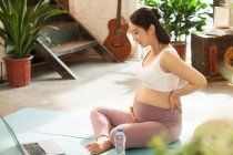 Улыбающаяся молодая беременная женщина сидит на ковриках для йоги и использует ноутбук дома — стоковое фото
