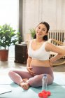 Молодая беременная азиатка сидит на ковриках для йоги и улыбается в камеру дома — стоковое фото