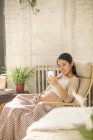 Lächelnde junge Schwangere sitzt im Schaukelstuhl und benutzt zu Hause ihr Smartphone — Stockfoto
