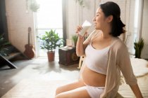 Feliz joven embarazada asiático mujer sentado y beber leche en casa - foto de stock
