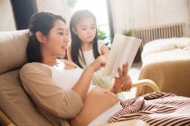 Jovem grávida leitura livro com adorável filhinha em casa — Fotografia de Stock