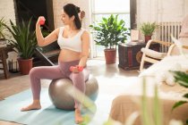 Jovem grávida sentada em forma de bola e exercitando-se com halteres em casa — Fotografia de Stock