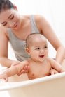 Happy young mother bathing adorable smiling infant baby sitting in bathtub — Stock Photo