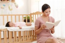 Giovane asiatico madre lettura libro mentre carino bambino dormire in presepe — Foto stock