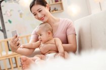 Sorrindo jovem mãe abraçando bebê e usando smartphone em casa — Fotografia de Stock