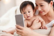 Усміхнена молода мати обіймає дитину і використовує смартфон — стокове фото